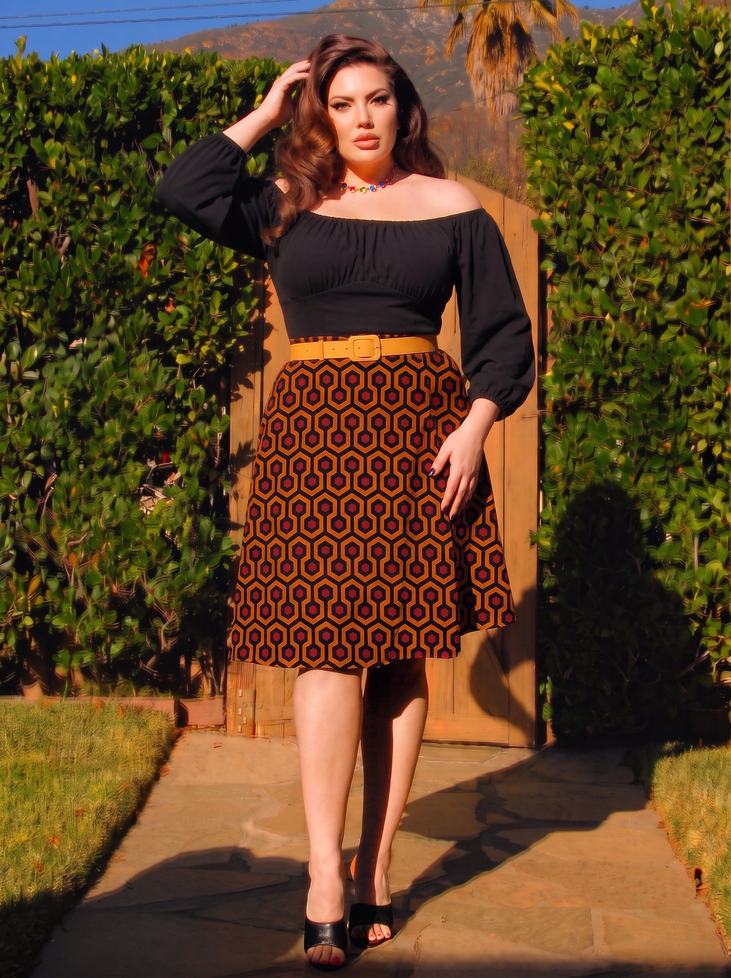 Full length view of the Viva 40s tulip skirt in orange and black Hotel Hexagon print, styled with a black off shoulder peasant top, a mustard belt and black peep toe heels