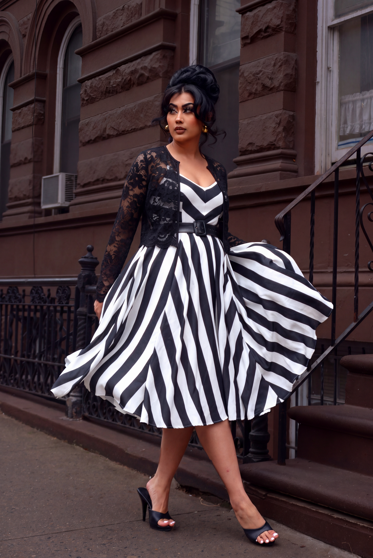Model wearing the Amalie 70s swing dress in black and white Mark Stripe crepe layered under a black sheer floral lace open front jacket, walking past a brownstone stoop