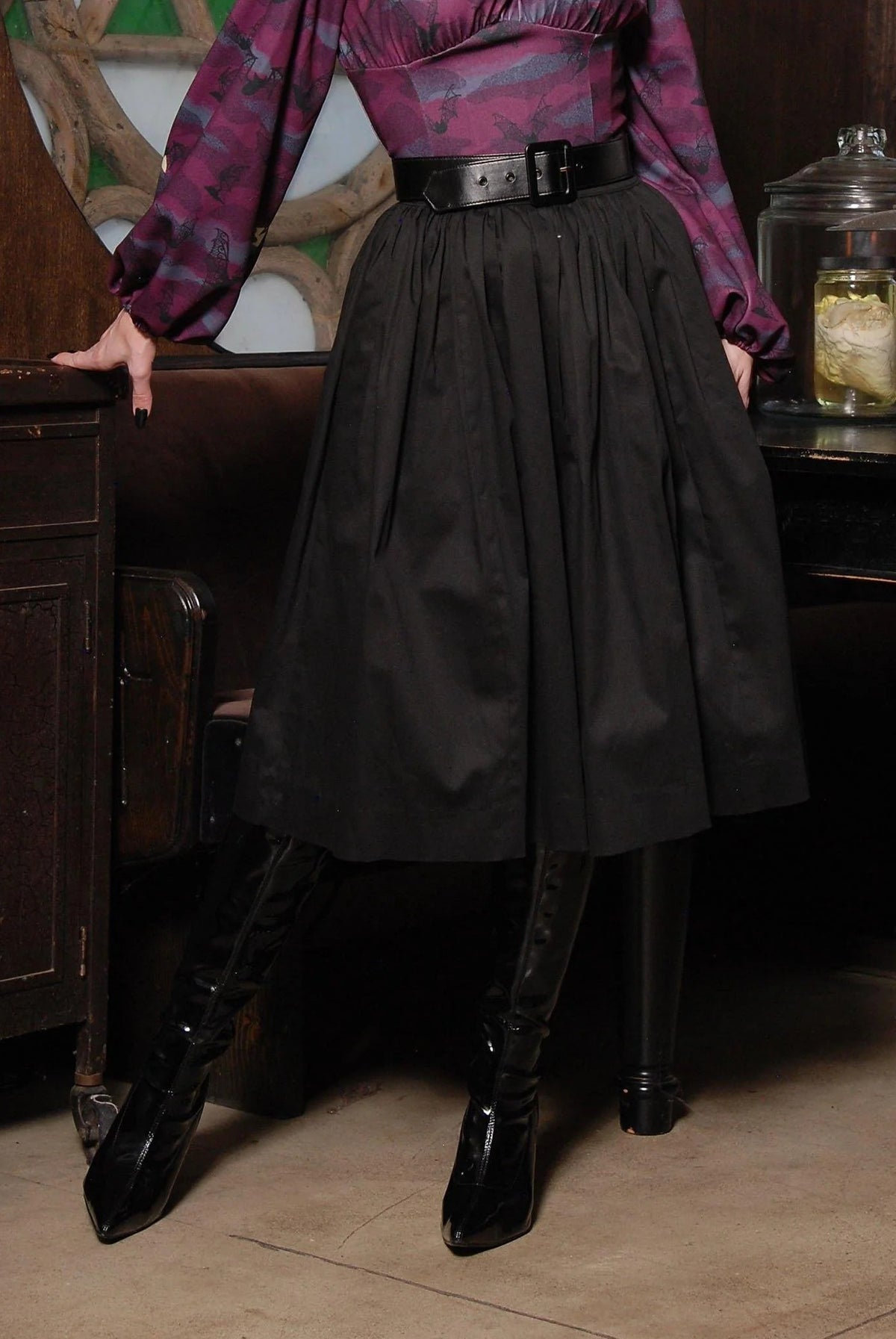 Waist-down view of the Bella gathered swing skirt in black cotton sateen with a black belt and black boots