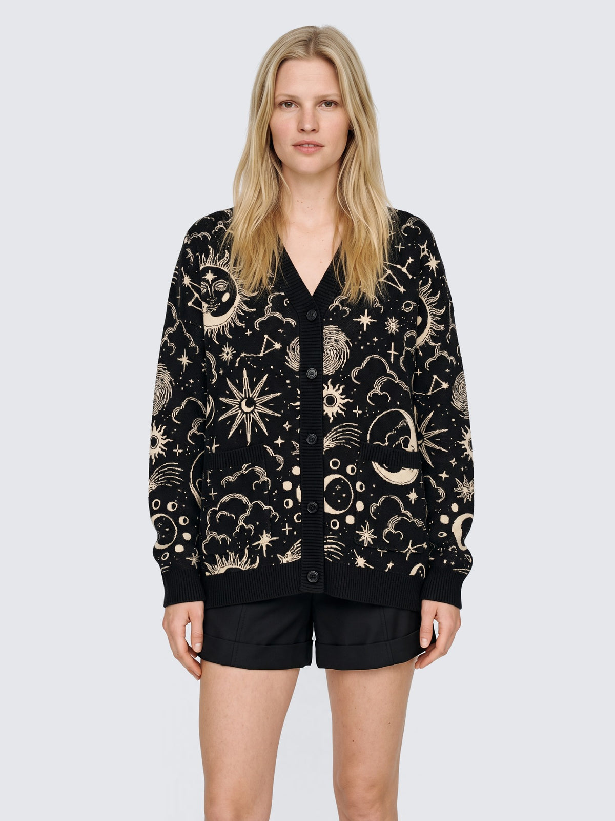 Model wearing the Celestial Cardigan oversized and buttoned, black knit with cream suns, moons and stars, with black high-waisted shorts