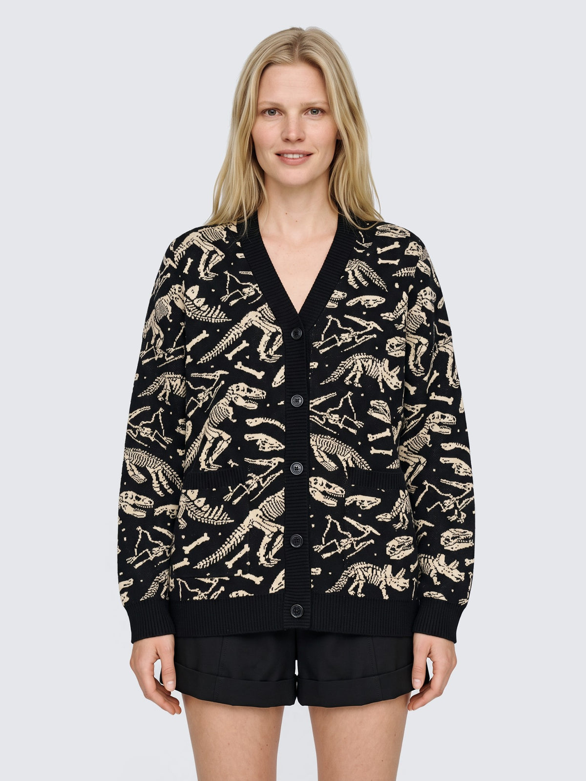 Model wearing the Dinosaur Fossil Cardigan oversized and buttoned, black knit with cream dinosaur skeletons, with black high-waisted shorts