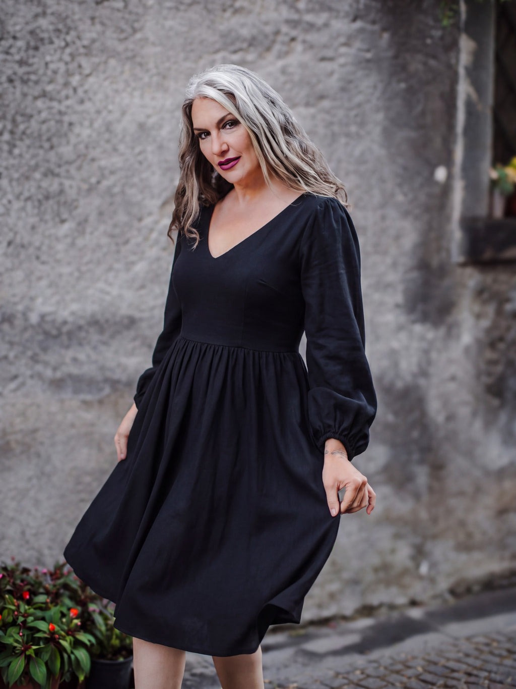 Kara swing dress in solid black against a stone wall in Sicily, with full billowing sleeves and a plunging V neckline, by Pinup Girl Clothing