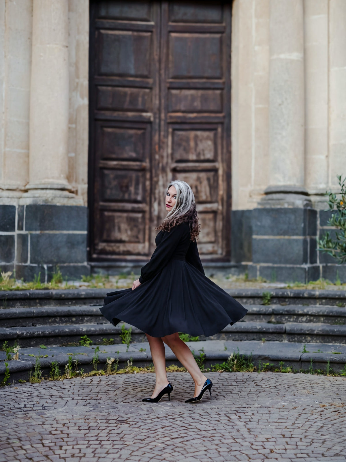 Kara swing dress in solid black twirling on cobblestones to show the full swing skirt, by Pinup Girl Clothing