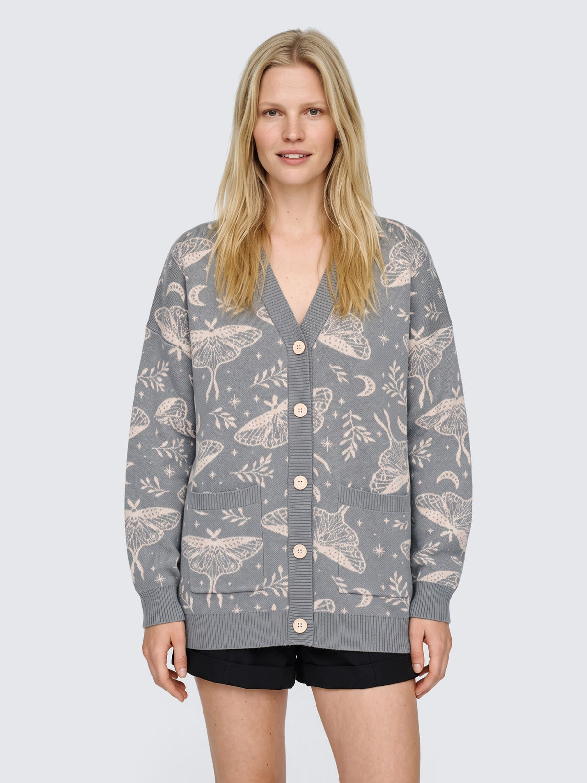 Model wearing the Luna Moth Cardigan oversized and buttoned, gray knit with cream luna moths and ferns, with black high-waisted shorts