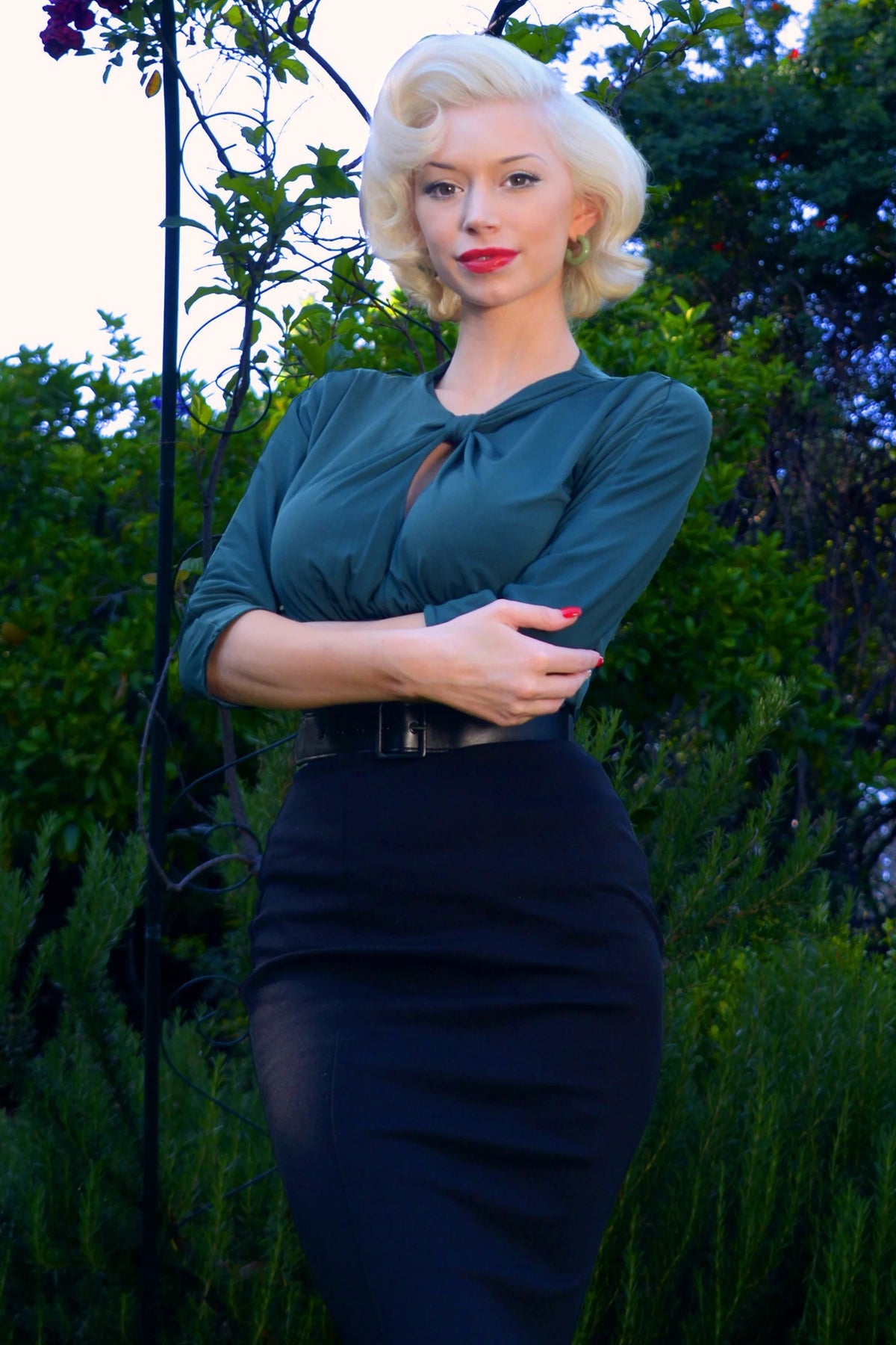 Model wearing the Malia top in dark green matte knit with its keyhole tie neckline and 3/4 sleeves, arms folded, with a navy pencil skirt in a garden