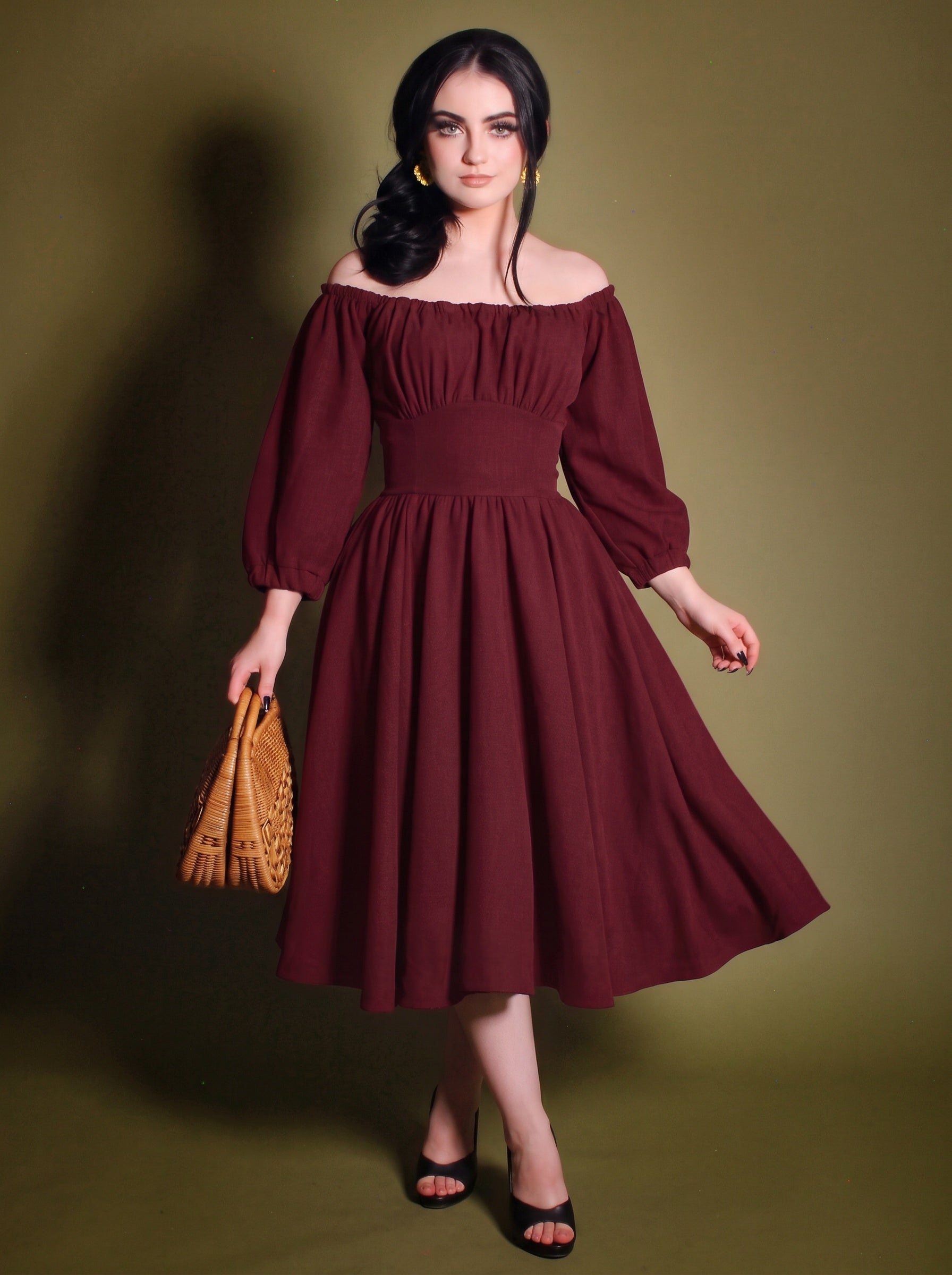 Model in the Marie-Therese burgundy peasant dress, a deep wine-red 70s day dress worn off the shoulder with long blouson sleeves, a wide fitted waistband and a full midi skirt with pockets