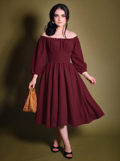 Model in the Marie-Therese burgundy peasant dress, a deep wine-red 70s day dress worn off the shoulder with long blouson sleeves, a wide fitted waistband and a full midi skirt with pockets
