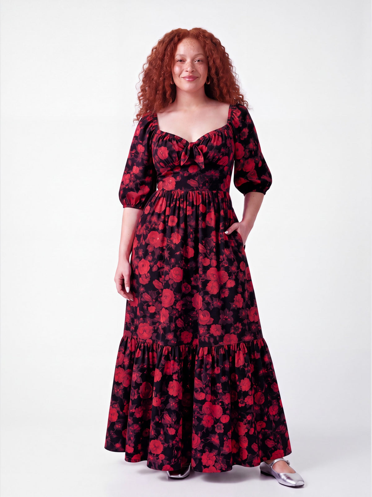 Model smiling softly in the Molly 70s Maxi Gown in Black and Red Ambrosia Floral, front view, one hand in the deep side pocket, tiered skirt to the floor