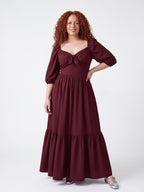 Model smiling in the Molly 70s Maxi Gown in Burgundy, one hand on her hip, the tiered burgundy crepe skirt falling to the floor