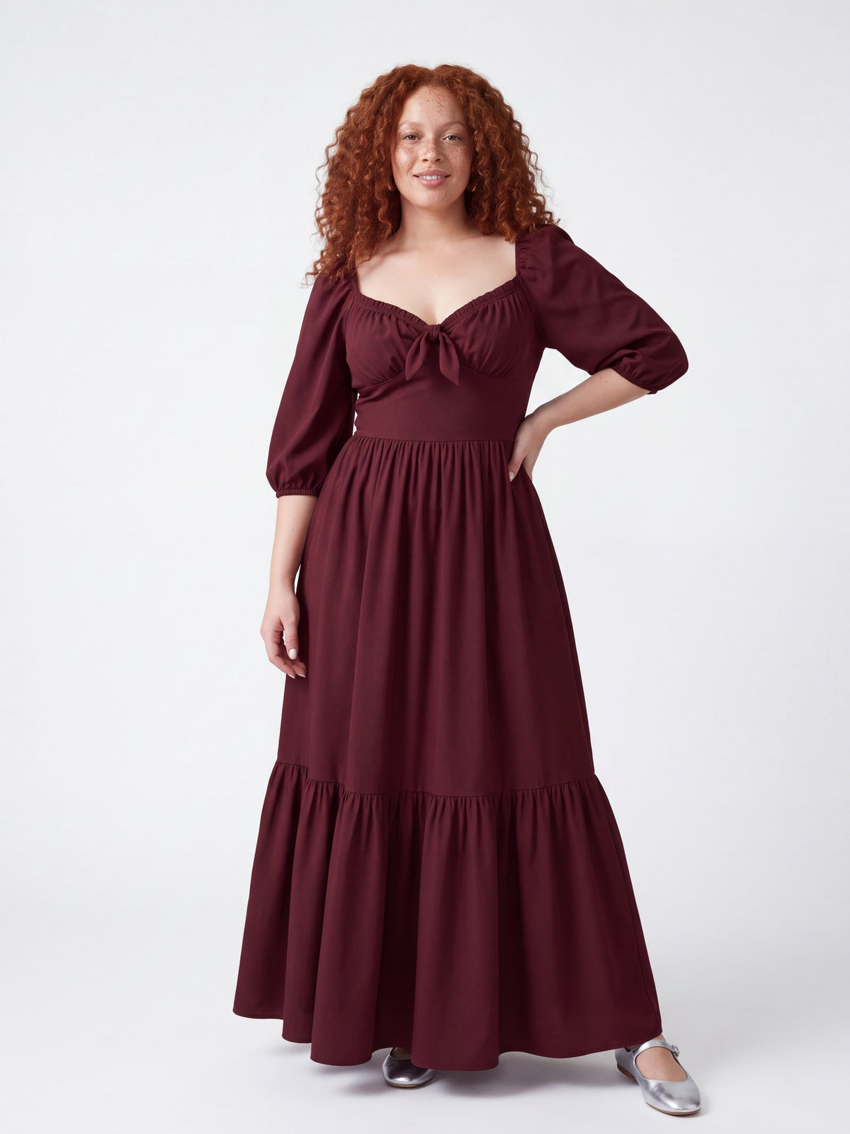 Model smiling in the Molly 70s Maxi Gown in Burgundy, one hand on her hip, the tiered burgundy crepe skirt falling to the floor
