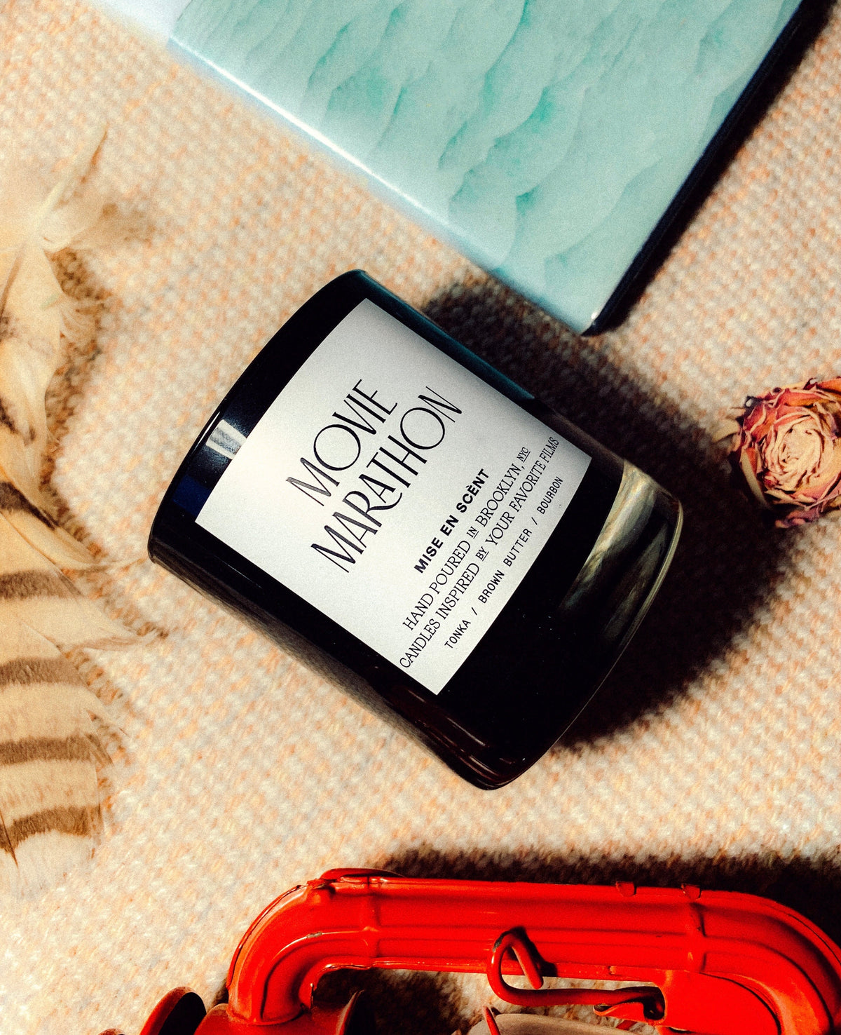 Movie Marathon candle lying on a cream blanket beside a red case