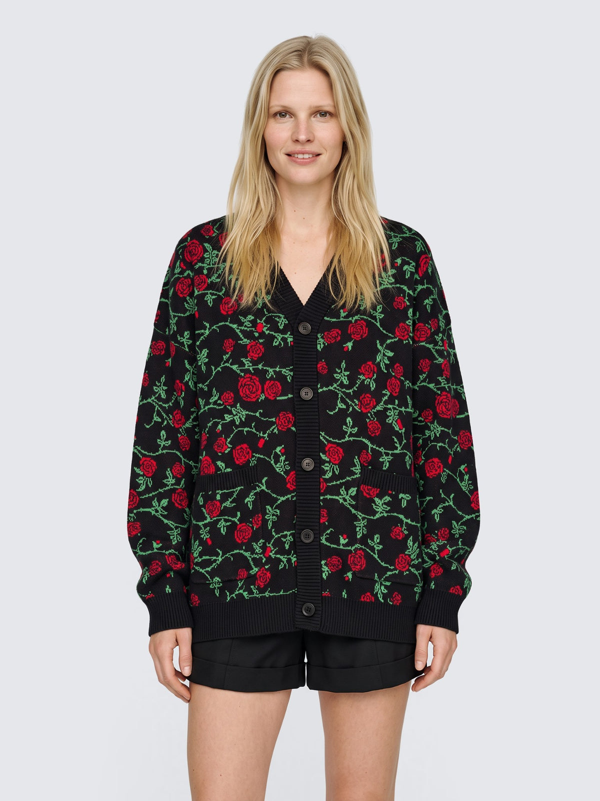Model wearing the Rose & Thorn Cardigan oversized and buttoned, black knit with red roses and green thorny vines, with black high-waisted shorts