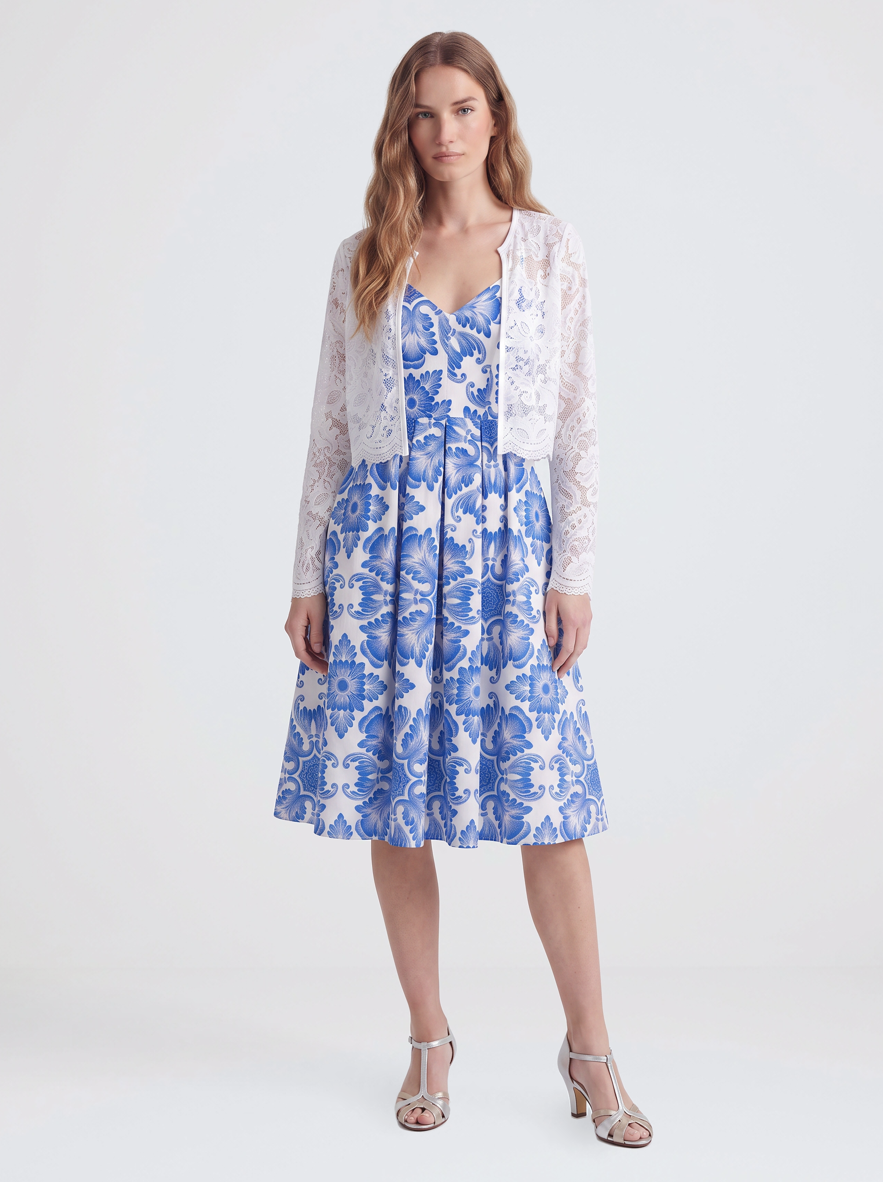 Model wearing the Amalie 70s ballerina swing dress in blue Noto tile print layered under a white sheer floral lace open front jacket, styled with silver T-strap heels