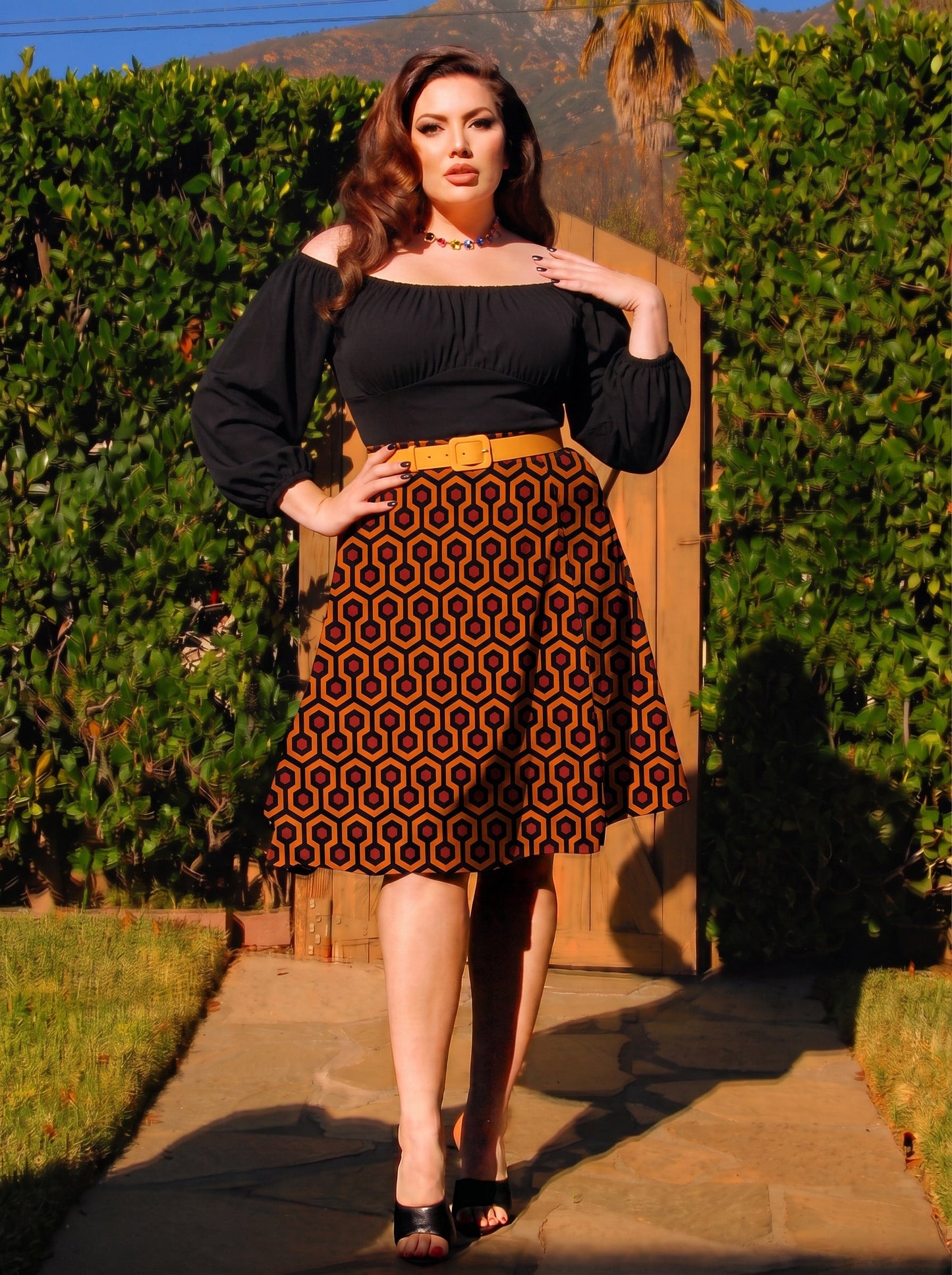 Model in the Viva 40s tulip skirt in orange and black Hotel Hexagon print, worn with a black peasant top and a mustard belt