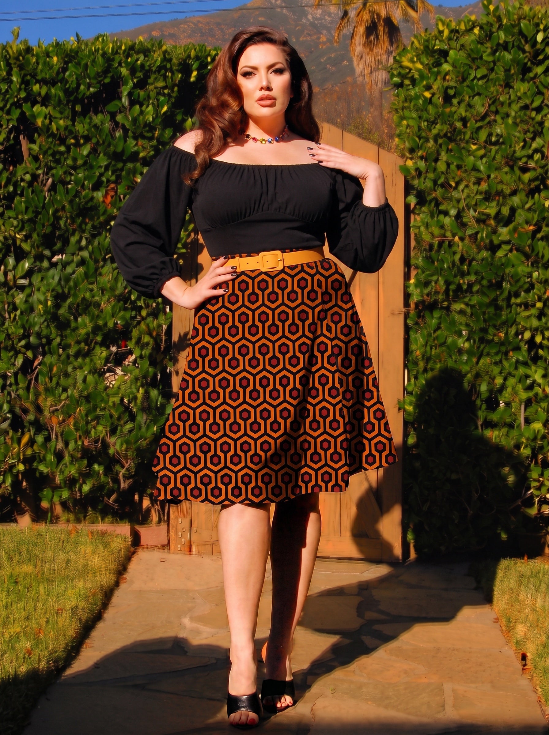 Model in the Viva 40s tulip skirt in orange and black Hotel Hexagon print, worn with a black peasant top and a mustard belt