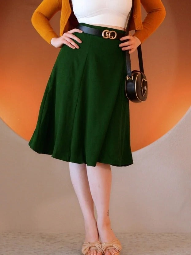 Front view of the Viva 40s Tulip Skirt in Hunter Green, high waisted with a black belt, flared skirt to just below the knee, hands on hips, worn with tan sandals