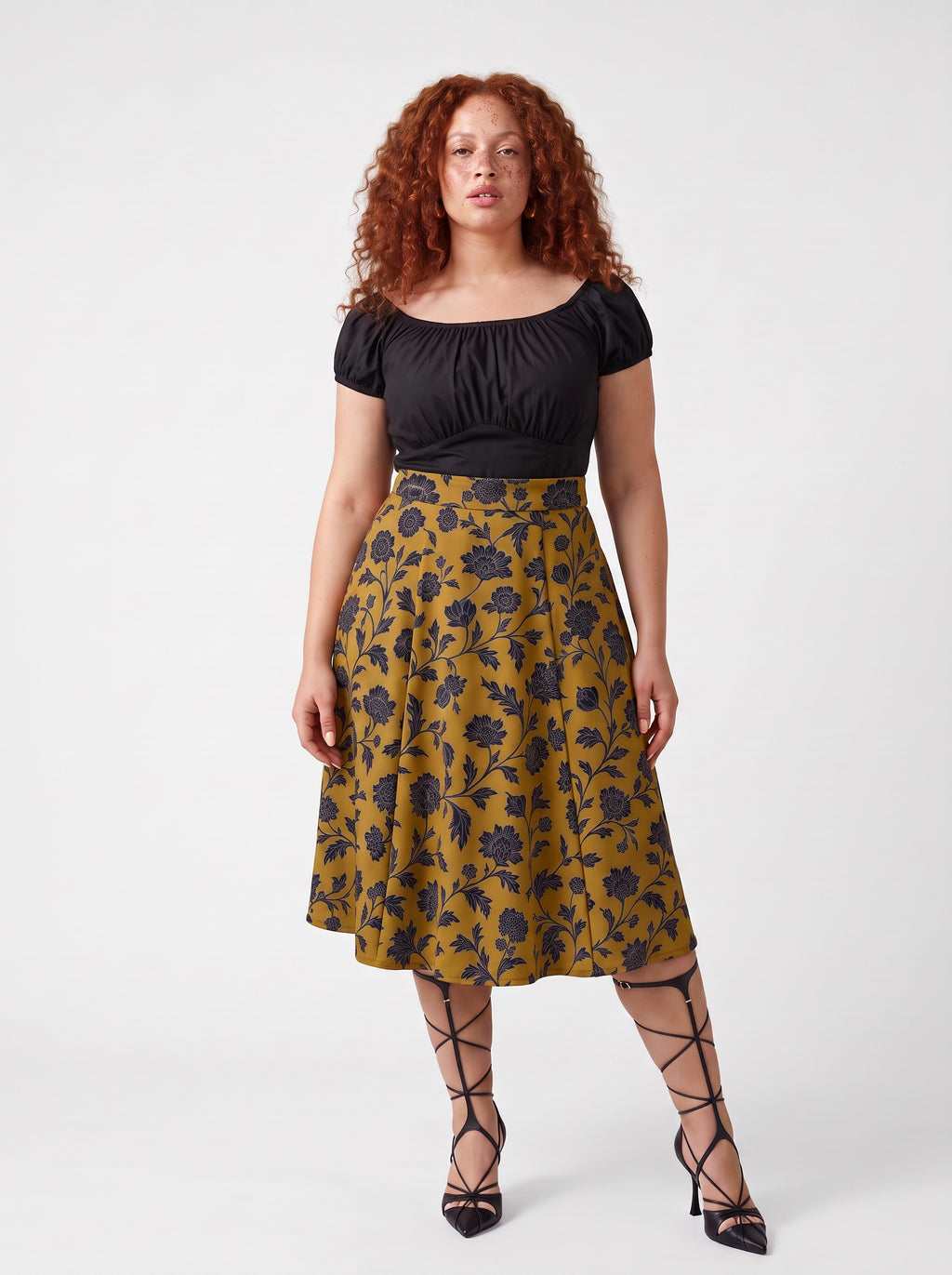 Model wearing the Viva 40s tulip swing skirt in ochre Capri Floral, black blooms on gold, with a black off shoulder peasant top and black heels