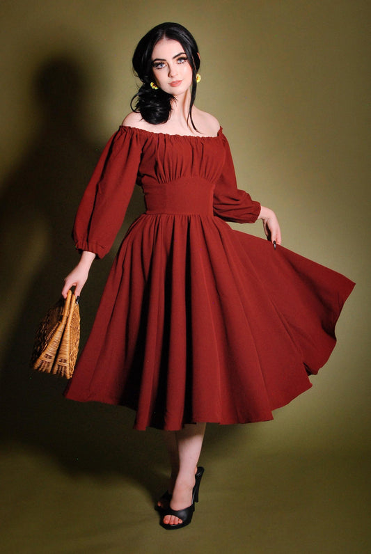 Model in the Marie-Therese merlot peasant dress, a deep wine-red 70s day dress worn off the shoulder with long blouson sleeves, a fitted waistband and a full twirl skirt with pockets
